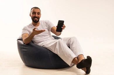 Pointing to a black screen on a phone, using modern technology devices, making video calls over the internet, a portrait of an Arab Saudi Gulf man wearing white pants and a t-shirt sitting on a comfortable chair holding a mobile phone, the concept of masculinity and self-confidence, white background.