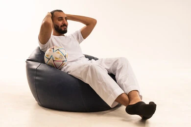 أنديه رياضية مختلفة، وضع اليدين خلف الرأس بايماءات الصدمة والاندهاش، إيماءات الشغف والتشجيع للفريق، تنمية الهوايات الرياضية، مشاهدة ومتابعة مباراة كرة القدم،  بورتريه لرجل عربي خليجي سعودي مبتسم يرتدي سروال وفانيلا  باللون الأبيض يجلس على كرسي مريح يحمل الكرة، خلفية بيضاء .