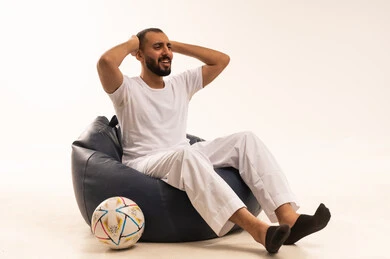 وضع اليدين خلف الرأس بايماءات الصدمة والاندهاش، إيماءات الشغف والتشجيع للفريق، تنمية الهوايات الرياضية، مشاهدة ومتابعة مباراة كرة القدم،  بورتريه لرجل عربي خليجي سعودي مبتسم يرتدي سروال وفانيلا باللون الأبيض يجلس على كرسي مريح يحمل الكرة، أنديه رياضية مختلفة، خلفية بيضاء .