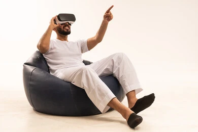 Attempting to click on something, a portrait of a Saudi Arabian Gulf man wearing white pants and a t-shirt sitting on a comfortable chair wearing 3D glasses, raising his index finger upwards, a young Saudi man pointing to a place with expressions of immersion and simulation of the imaginary world, white background.