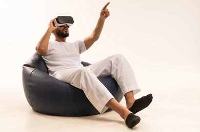 A Saudi young man points to a place with expressions of immersion and simulation of the imaginary world, trying to click on something. A portrait of an Arab Gulf Saudi man wearing a white suit sitting on a comfortable chair, wearing 3D glasses, raising his index finger upwards, with a white background.