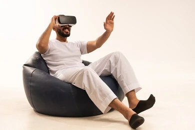 Interaction, integration, and simulation of the imaginary world, an attempt to click on something, using modern technology and devices, a portrait of a Saudi Arabian Gulf man wearing white pants and a t-shirt sitting on a comfortable chair wearing 3D glasses raising his index finger upwards, white background.