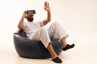 Gestures of enthusiasm and engagement, interaction and simulation of the imaginary world, an attempt to click on something, using modern technology and devices, a portrait of an Arab Gulf Saudi man wearing white pants and a t-shirt sitting on a comfortable chair wearing 3D glasses raising his index finger upwards, white background.