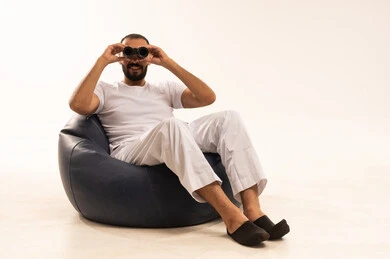 حب الاستكشاف والمغامرة، رؤية الاشياء البعيدة و تقريبها, التخطيط المستقبلي للأهداف والطموحات,  أنشطة وفعاليات مسلية،  بورتريه لرجل عربي سعودي خليجي يرتدي سروال وفانيلا أبيض يجلس على كرسي مريح ويضع على عيونه منظار، خلفية بيضاء