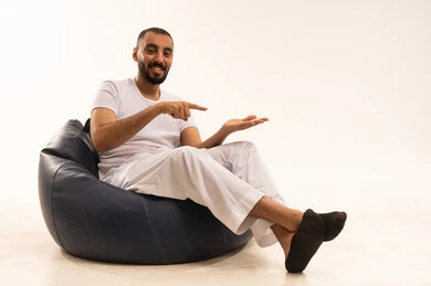 Lifting and carrying something, indicating and clarifying something, specifying a certain location, pointing to the place of an element, elegance and attention to appearance, a portrait from behind of an Arab Saudi Gulf man wearing white pants and a t-shirt sitting on a comfortable chair and pointing with his hand, white background.