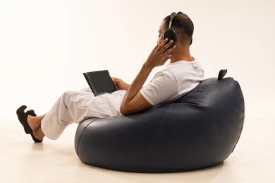 Watching programs and videos using a tablet, using modern technology devices, completing daily tasks remotely, a portrait from behind of an Arab Saudi Gulf man wearing white pants and a t-shirt sitting on a comfortable chair and wearing headphones, the concept of masculinity and self-confidence, white background.