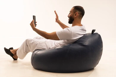 Video call using a mobile device, using modern technology devices, completing daily tasks remotely, a portrait from behind of an Arab Saudi Gulf man wearing white pants and a t-shirt sitting on a comfortable chair holding a mobile phone, the concept of masculinity and self-confidence, white background.