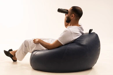 استعمال المنظار للكشف عن الاشياء البعيدة وتقريبها, حب الاستكشاف والمغامرة، رؤية الاشياء البعيدة و تقريبها, التخطيط المستقبلي للأهداف والطموحات, بورتريه من الخلف لرجل عربي سعودي خليجي يرتدي سروال وفانيلا أبيض يجلس على كرسي مريح ويضع على عيونه منظار، خلفية بيضاء