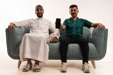 شاشة سوداء على هاتف محمول، التفاعل بمشاهدة الفريق المفضل, ايماءات التحدي والانتصار، قضاء اوقات الفراغ الممتعة مع الاصدقاء، أنشطة رياضية ترفيهية،  بورتريه لرجلين عربيين خليجيين سعوديين احدهما يمسك بيده كرة، شاب سعودي يرتدي قميص المنتخب السعوديي، خلفية بيضاء
