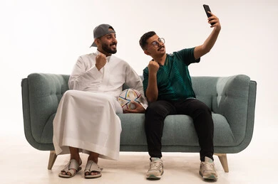 A selfie to document moments, interacting while watching the favorite team, gestures of challenge and victory, spending enjoyable free time with friends, recreational sports activities, a portrait of two Arab Gulf Saudi men, one holding a ball, a Saudi young man wearing the Saudi national team jersey, white background.