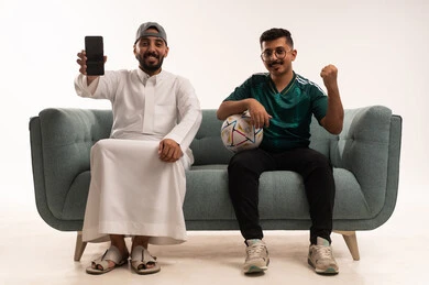الجهاز المحمول يعرض شاشة فارغة سوداء، التفاعل بمشاهدة الفريق المفضل, ايماءات التحدي والانتصار، قضاء اوقات الفراغ الممتعة مع الاصدقاء، أنشطة رياضية ترفيهية،  بورتريه لرجلين عربيين خليجيين سعوديين احدهما، شاب سعودي يرتدي قميص المنتخب السعوديي، خلفية بيضاء