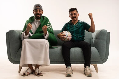 Spending enjoyable leisure time with friends, excitement and interaction with the national team, recreational sports activities, passion for following the national team, a portrait of two Arab Gulf men from Saudi Arabia, one of whom is draping the flag of the Kingdom over his shoulders, a young Saudi man wearing the Saudi national team jersey and holding a ball, white background.