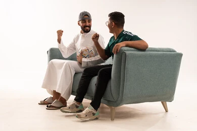 الشغف في متابعة فريق المنتخب الوطني، أنشطة رياضية ترفيهية، إيماءات الشغف والتشجيع، مفهوم الفوز و الانتصار في المباريات،  بورتريه من الجانب لرجلين عربيين خليجيين سعوديين احدهما يحمل كرة، الجلوس على آريكة مريحة، شاب سعودي يرتدي قميص المنتخب السعوديي، خلفية بيضاء