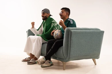 مشاهدة المنتخب الوطني، أنشطة رياضية ترفيهية، إيماءات الشغف والتشجيع، مفهوم الفوز و الانتصار في المباريات،  بورتريه من الجانب لرجلين عربيين خليجيين سعوديين احداهما يضع على كتفه علم المملكة، الجلوس على آريكة مريحة، شاب سعودي يرتدي قميص المنتخب السعوديي، خلفية بيضاء