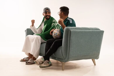 أنشطة رياضية ترفيهية، إيماءات الشغف والتشجيع، مفهوم الفوز و الانتصار في المباريات، مشاهدة المنتخب الوطني، بورتريه من الجانب لرجلين عربيين خليجيين سعوديين احداهما يضع على كتفه علم المملكة، الجلوس على آريكة مريحة، شاب سعودي يرتدي قميص المنتخب السعوديي، خلفية بيضاء