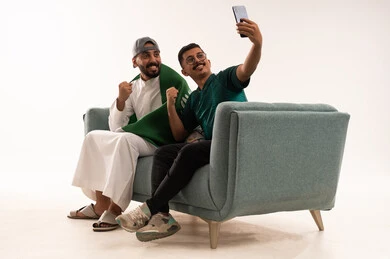 التقاط صورة مع حركات التشجيع، التفاعل بمشاهدة الفريق المفضل, ايماءات التحدي والانتصار، قضاء اوقات الفراغ الممتعة مع الاصدقاء، أنشطة رياضية ترفيهية،  بورتريه لرجلين عربيين خليجيين سعوديين احدهما، شاب سعودي يرتدي قميص المنتخب السعوديي، خلفية بيضاء