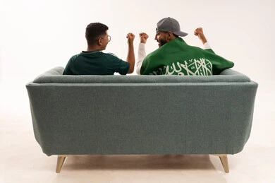 Gestures of joy and happiness, the concept of winning and victory in matches, recreational sports activities, the feeling of winning and triumph, a portrait from behind of two Arab Gulf Saudi men, one of whom has the flag of the Kingdom draped over his shoulder, sitting on a comfortable sofa, a young Saudi man wearing the Saudi national team jersey, white background.