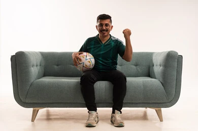 رفع قبضة اليد بإيماءات الفوز، تنمية الهوايات الرياضية، مشاهدة ومتابعة مباراة كرة القدم ،  إيماءات الشغف والتشجيع للفريق ، بورتريه لشاب عربي خليجي سعودي مبتسم يرتدي الزي الرياضي يجلس على اريكة مريحة يحمل الكرة ، الحماس والتفاعل مع المنتخب ، خلفية بيضاء .