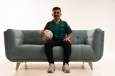 Developing sports hobbies, watching and following a football match, gestures of passion and encouragement for the team, a portrait of a smiling young Arab Gulf Saudi man wearing sportswear sitting on a comfortable sofa holding a ball, enthusiasm and interaction with the national team, white background.