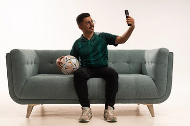 Video call on the phone while watching matches, interacting while watching the favorite team, gestures of challenge and victory, spending enjoyable free time with friends, recreational sports activities, a portrait of a young Saudi man wearing the Saudi national team jersey and holding a ball in his hand, white background.