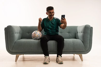 مشاهدة المباريات على الهاتف المحمول، دمج التقنيات مع الحياة اليومية، التفاعل بمشاهدة الفريق المفضل, ايماءات التحدي والانتصار، قضاء اوقات الفراغ الممتعة مع الاصدقاء، أنشطة رياضية ترفيهية،  بورتريه لشاب سعودي يرتدي قميص المنتخب السعوديي ويحمل بيده كرة، خلفية بيضاء