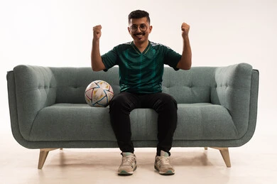 رفع قبضة اليد بإيماءات الفوز، تنمية الهوايات الرياضية، مشاهدة ومتابعة مباراة كرة القدم ،  إيماءات الشغف والتشجيع للفريق ، بورتريه لشاب عربي خليجي سعودي مبتسم يرتدي الزي الرياضي يجلس على اريكة مريحة يحمل الكرة ، الحماس والتفاعل مع المنتخب ، خلفية بيضاء .