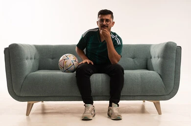 النظر إلى شيء ما، تنمية الهوايات الرياضية، مشاهدة ومتابعة مباراة كرة القدم ،  إيماءات الشغف والتشجيع للفريق ، بورتريه لشاب عربي خليجي سعودي مبتسم يرتدي الزي الرياضي يجلس على اريكة مريحة يحمل الكرة ، الحماس والتفاعل مع المنتخب ، خلفية بيضاء .