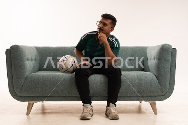 النظر إلى شيء ما، تنمية الهوايات الرياضية، مشاهدة ومتابعة مباراة كرة القدم ،  إيماءات الشغف والتشجيع للفريق ، بورتريه لشاب عربي خليجي سعودي مبتسم يرتدي الزي الرياضي يجلس على اريكة مريحة يحمل الكرة ، الحماس والتفاعل مع المنتخب ، خلفية بيضاء .