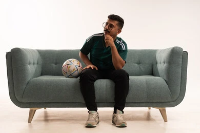 النظر إلى شيء ما، تنمية الهوايات الرياضية، مشاهدة ومتابعة مباراة كرة القدم ،  إيماءات الشغف والتشجيع للفريق ، بورتريه لشاب عربي خليجي سعودي مبتسم يرتدي الزي الرياضي يجلس على اريكة مريحة يحمل الكرة ، الحماس والتفاعل مع المنتخب ، خلفية بيضاء .