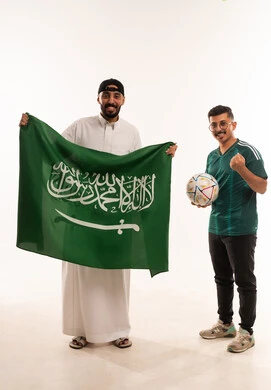 مفهوم الفوز و الانتصار في المباريات، أنشطة رياضية ترفيهية، إيماءات الفرح و السعادة، الشعور بالفرح والسرور، بورتريه  لرجلين عربيين خليجيين سعوديين احداهما يحمل كرة القدم والآخر يحمل علم المملكة، شاب سعودي يرتدي قميص المنتخب السعوديي لكررة القدم، خلفية بيضاء