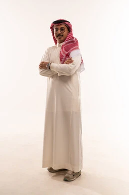 الوقوف بأيدي متقاطعة والنظر إلى الكاميرا بتعابير السرور، بورتريه لشاب عربي خليجي سعودي يرتدي الثوب والشماغ التقليدي ويضع النظارة الطبية، الاناقة والاهتمام بالمظهر الخارجي، الطول الكامل للجسم، خلفية بيضاء