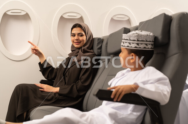 رحلة سياحية عائلية، امرأة عربية خليجية عمانية ترتدي الحجاب و العباءة برفقة ولدها في الطائرة، الاشارة الي شيء ما، قضاء العطلة الصيفية برفقة العائلة، خطوط الطيران العماني ايماءات السعادة و السرور