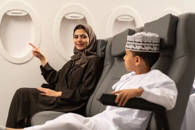 A family tourist trip, an Arab Gulf Omani woman wearing a hijab and abaya with her son on the plane, pointing to something, spending the summer vacation with family, Oman Air gestures of happiness and joy.