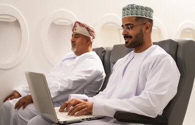 العمل لتحقيق الاهداف والاستراتيجيات, إدارة شؤون الأعمال وتنظيمها عن بعد،  رجل عربي خليجي عماني يرتدي الدشداشة و الكمة يجلس داخل الطائرة فى رحله جوية  برفقة والده المسن يقوم بالعمل على الحاسوب المحمول , استخدام الأجهزة التكنولوجية الحديثة والمتطورة , مفهوم السياحه و السفر