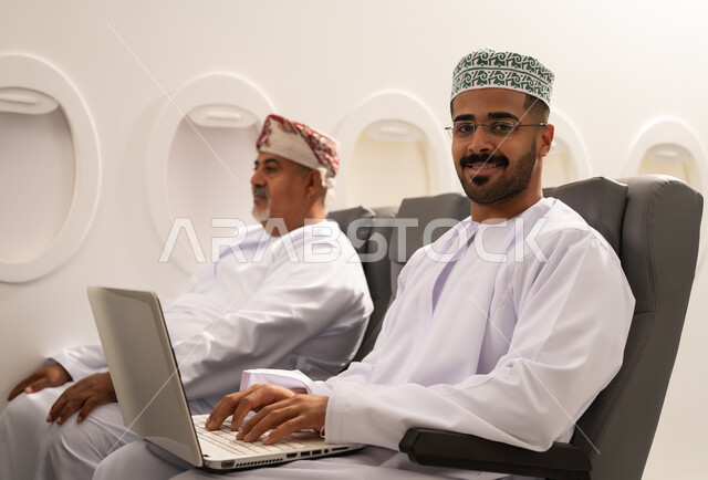 النظر الى الكاميرا بايماءات السعادة و السرور, إدارة شؤون الأعمال وتنظيمها عن بعد،  رجل عربي خليجي عماني يرتدي الدشداشة و الكمه يجلس داخل الطائرة فى رحله جوية  برفقة والده المسن يقوم بالعمل على الحاسوب المحمول , استخدام الأجهزة التكنولوجية الحديثة والمتطورة , مفهوم السياحه و السفر
