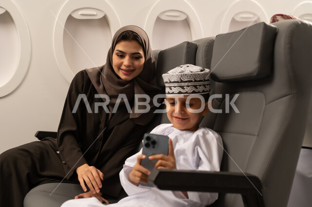 قضاء الأوقات الممتعة مع الأبناء,توثيق اللحظات المميزة باستخدام الأجهزة التقنية الحديثة, امرأة عربية خليجية إماراتية مع ابنها داخل الطائرة فى رحله جوية يرتديان الزي التقليدي الماني يقومان بالتقاط صور السيلفي التذكارية عبر الجوال, الأجواء العائلية السعيدة, مفهوم السياحة و السفر