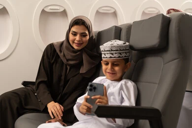 قضاء الأوقات الممتعة مع الأبناء,توثيق اللحظات المميزة باستخدام الأجهزة التقنية الحديثة, امرأة عربية خليجية إماراتية مع ابنها داخل الطائرة فى رحله جوية يرتديان الزي التقليدي الماني يقومان بالتقاط صور السيلفي التذكارية عبر الجوال, الأجواء العائلية السعيدة, مفهوم السياحة و السفر