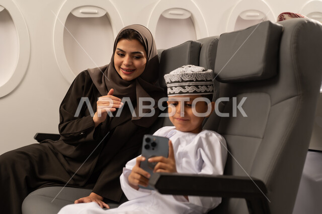 توثيق اللحظات المميزة باستخدام الأجهزة التقنية الحديثة, قضاء الأوقات الممتعة مع الأبناء, امرأة عربية خليجية إماراتية مع ابنها داخل الطائرة فى رحله جوية يرتديان الزي التقليدي الماني يقومان بالتقاط صور السيلفي التذكارية عبر الجوال, الأجواء العائلية السعيدة, مفهوم السياحة و السفر