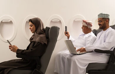 Browsing social media, using a modern and advanced technological device in daily life, chatting with family and friends, an Omani Gulf Arab woman wearing a hijab and abaya sitting inside an airplane on a flight, an elderly Gulf man wearing a dishdasha and ghutrah taking a selfie with a mobile phone, the concept of tourism and travel.