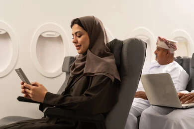 The use of modern and advanced technology devices in daily life, browsing social media, chatting with family and friends, an Arab Gulf Omani woman wearing a hijab and abaya sitting inside an airplane on a flight, the concept of tourism and travel, gestures of happiness and joy.