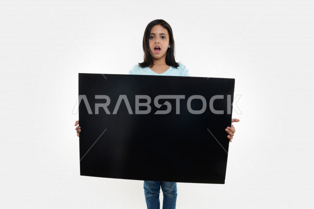 بورتريه لفتاة عربية خليجية سعودية تحمل بيديها لوحة سوداء فارغة بإيماءات تدل على الدهشة ، لوحة إعلانية ، لوحة فنية ، خلفية بيضاء