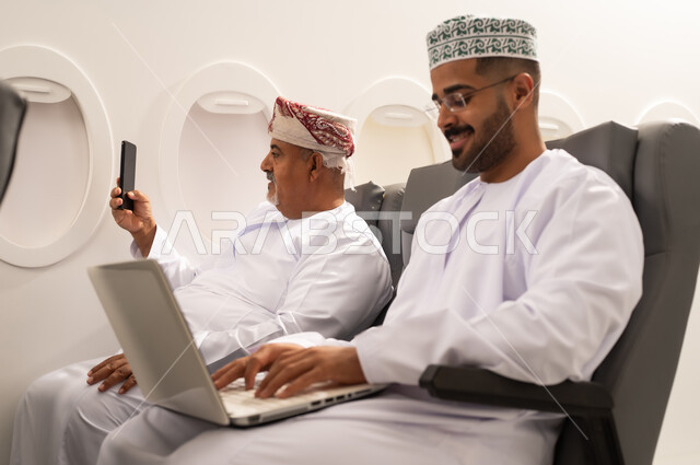 إنجاز المهام المطلوبة عبر اللابتوب, إلتقاط صورة سيلفي من خلال الهاتف المحمول, ادارة الأعمال عن بعد، استخدام الأجهزة الحديثة والمتطورة، رجل عربية خليجي عماني يرتدي الدشداشة و الكمه يجلس داخل الطائرة فى رحله جوية برفقة والده المسن يقوم باستخدام الحاسوب المحمول، دمج التكنولوجيا بالعمل, مفهوم السياحة و السفر