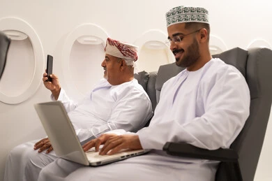 إنجاز المهام المطلوبة عبر اللابتوب, إلتقاط صورة سيلفي من خلال الهاتف المحمول, ادارة الأعمال عن بعد، استخدام الأجهزة الحديثة والمتطورة، رجل عربية خليجي عماني يرتدي الدشداشة و الكمه يجلس داخل الطائرة فى رحله جوية برفقة والده المسن يقوم باستخدام الحاسوب المحمول، دمج التكنولوجيا بالعمل, مفهوم السياحة و السفر