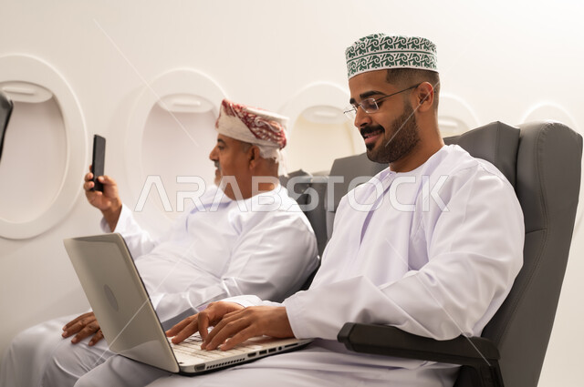 إلتقاط صورة سيلفي من خلال الهاتف المحمول, ادارة الأعمال عن بعد، استخدام الأجهزة الحديثة والمتطورة، رجل عربية خليجي عماني يرتدي الدشداشة و الكمه يجلس داخل الطائرة فى رحله جوية برفقة والده المسن يقوم باستخدام الحاسوب المحمول، دمج التكنولوجيا بالعمل، إنجاز المهام المطلوبة عبر اللابتوب, مفهوم السياحة و السفر