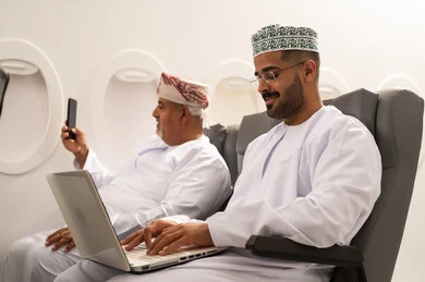 إلتقاط صورة سيلفي من خلال الهاتف المحمول, ادارة الأعمال عن بعد، استخدام الأجهزة الحديثة والمتطورة، رجل عربية خليجي عماني يرتدي الدشداشة و الكمه يجلس داخل الطائرة فى رحله جوية برفقة والده المسن يقوم باستخدام الحاسوب المحمول، دمج التكنولوجيا بالعمل، إنجاز المهام المطلوبة عبر اللابتوب, مفهوم السياحة و السفر