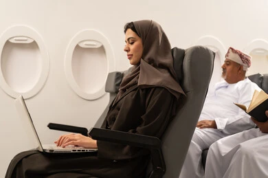 The use of modern and advanced technology, accomplishing remote work tasks through the internet, making video calls via a laptop, an Arab Gulf Omani young woman wearing an abaya and hijab sitting inside an airplane on a flight working on a laptop, the concept of tourism and travel.