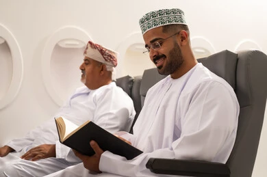 The culture of self-learning, spending free time on learning and culture, practicing the hobby of reading, an Arab Gulf Omani man wearing a dishdasha and kumma sitting inside an airplane on a flight with his elderly father, the concept of tourism and travel.