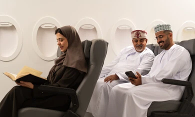 Learning and reading during free time, watching movies and plays on the mobile, two Arab Gulf men from Oman wearing the dishdasha and kumma sitting inside the airplane on a flight, a Gulf woman reading a book, gestures of happiness and joy, the concept of tourism and travel.
