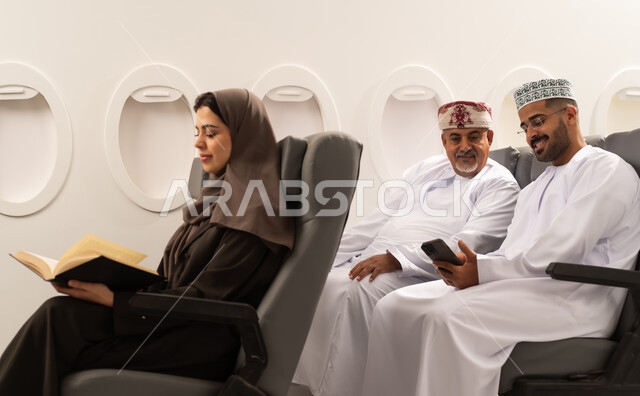الاستمتاع بالقراءة وتوسيع المعرفة,مشاهده الافلام و المسرحيات عبر الجوال , رجلان عربيان خليجيان عمانيان يرتديان الدشداشة و الكمه يجلسان داخل الطائرة فى رحله جوية , امراة خليجية تقوم بقراءة كتاب,  ايماءات السعادة و السرور , مفهوم السياحه و السفر