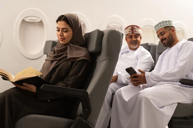 Spending time reading, learning, and gaining knowledge, an Omani Gulf Arab family dressed in traditional attire sitting inside the airplane on a tourism trip, two Gulf men watching a mobile phone, a Gulf woman holding a book in her hands, the concept of tourism and travel.
